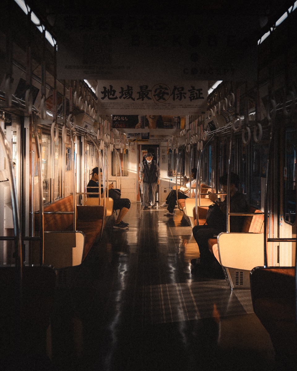 A lone figure stands in a warm-lit train carriage at night — the observer's view Jun shoots from.