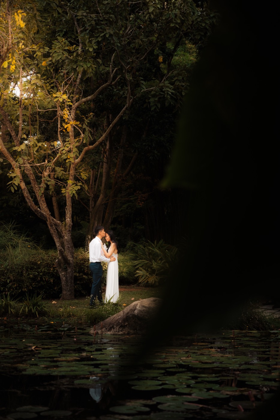A couple stand close at the water's edge in the last blue light, framed by dark foliage and a still lily pond.