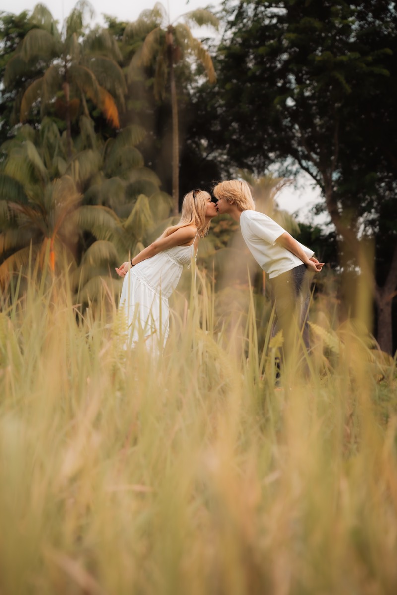 A couple lean in for a kiss amid soft, sunlit grass.