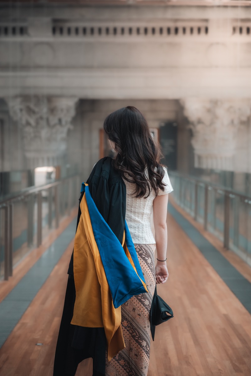 A graduate seen from behind, gown over one shoulder, walking a grand pillared hall.
