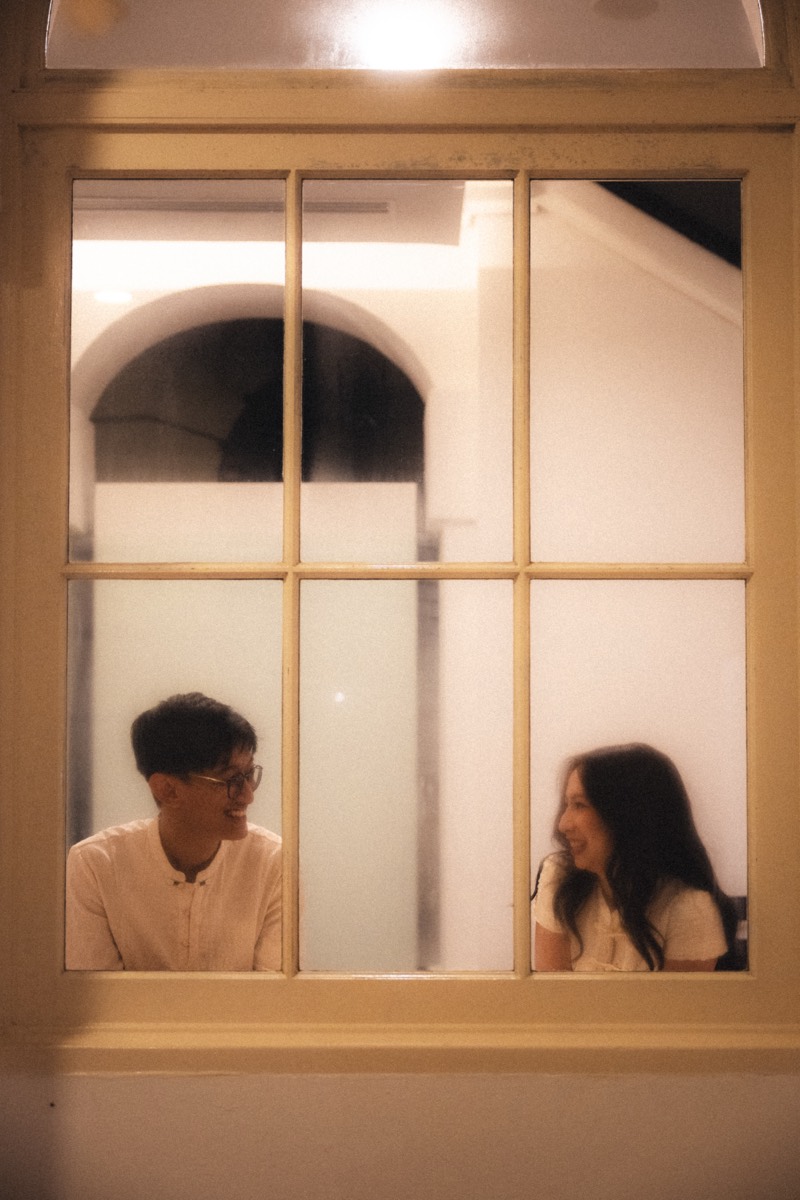 A couple framed in a lamplit window at night, mid-conversation.