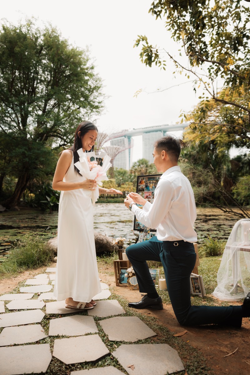 A man kneels to propose on a garden path by the water, his partner holding a bouquet.