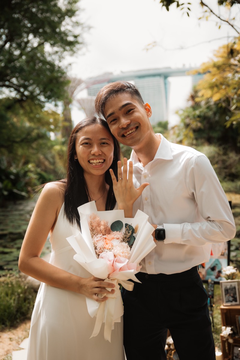 A newly engaged couple smile together, showing the ring, Marina Bay Sands behind them.