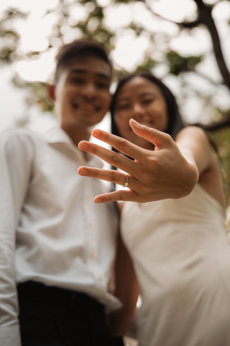 The couple hold the ring up to the camera, laughing.