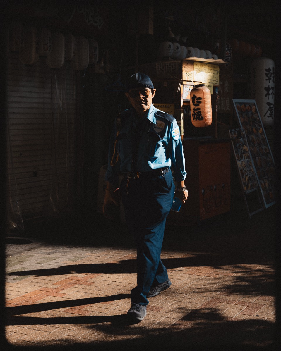 A lone figure walks through a shaft of light past shuttered shopfronts and paper lanterns.