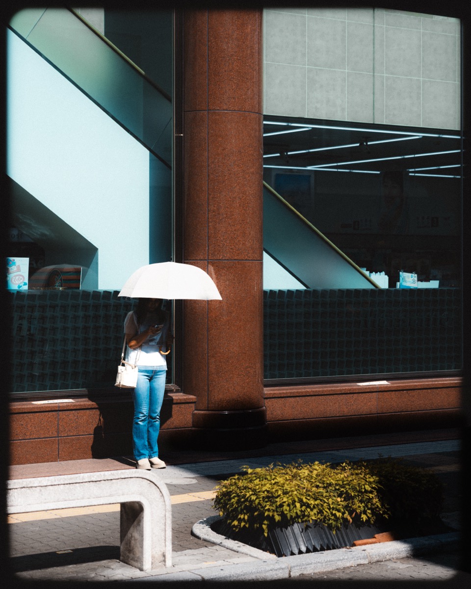 A person under a white umbrella waits beside a column in teal and red shadow.
