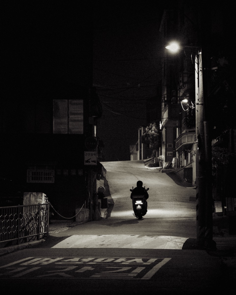 A lone scooter rider rolls down a quiet sloped alley under a single streetlight.