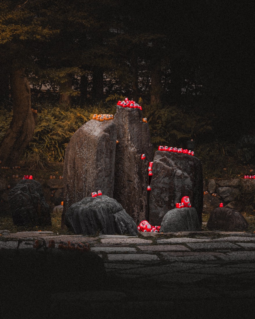 Offerings — red offerings and candles glow among dark stones at a forest shrine.