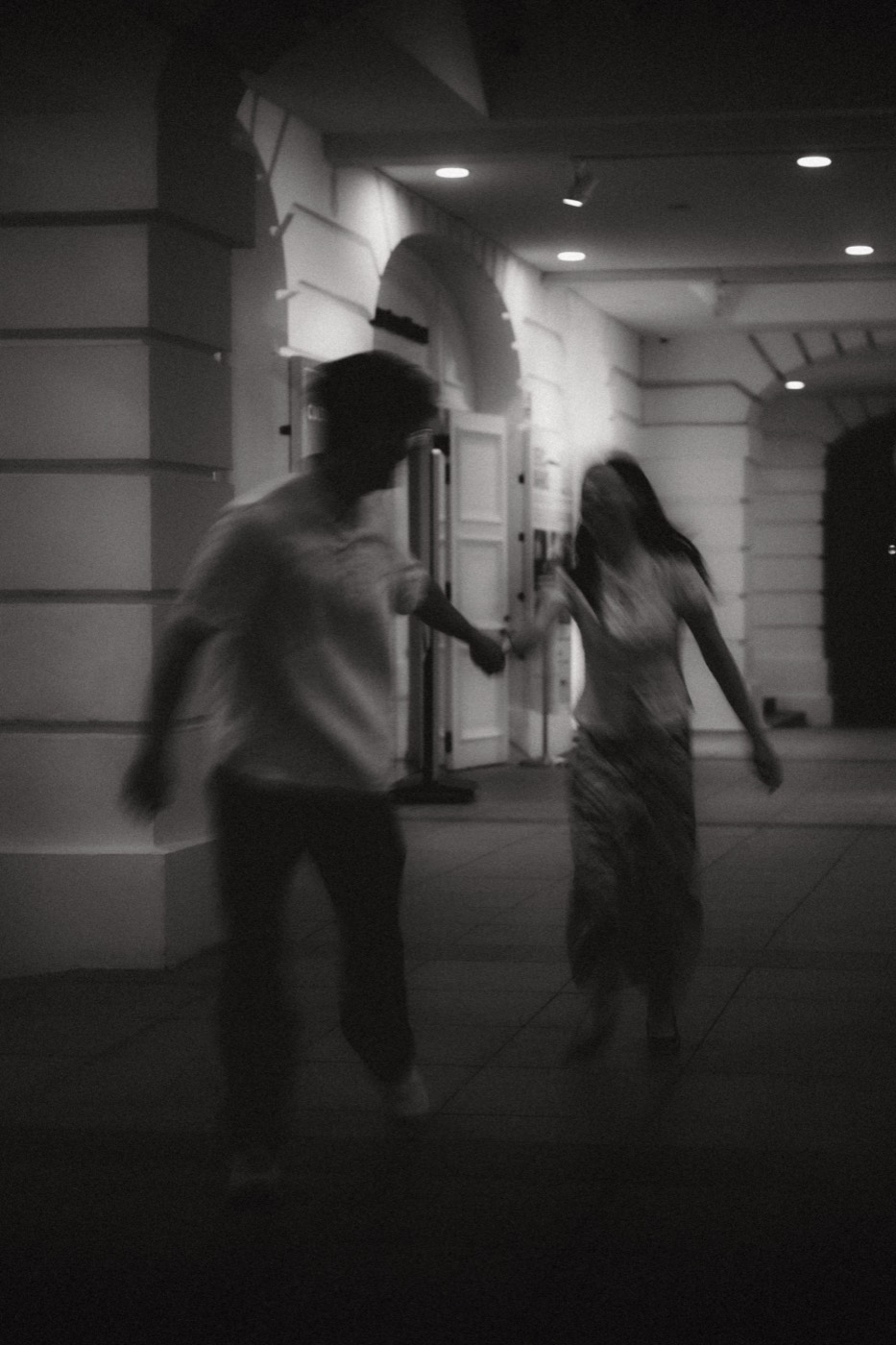 A couple run hand in hand through lamplit colonial arches, caught mid-motion in black and white.